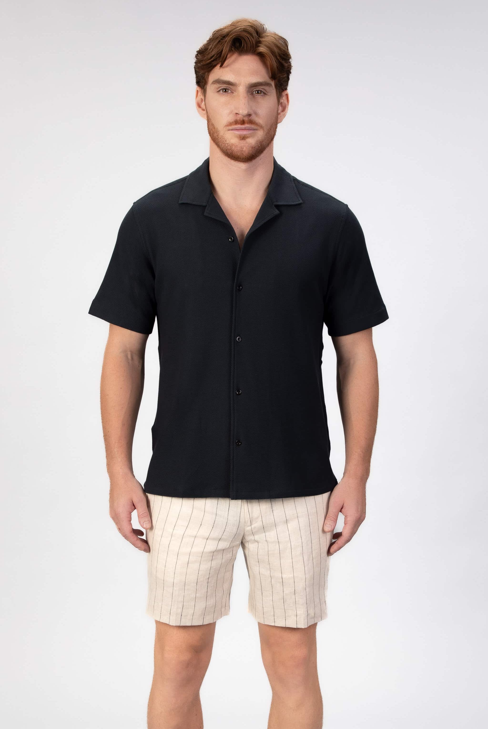 Man wearing a black short-sleeve camp collar shirt and beige stripe shorts on a plain background