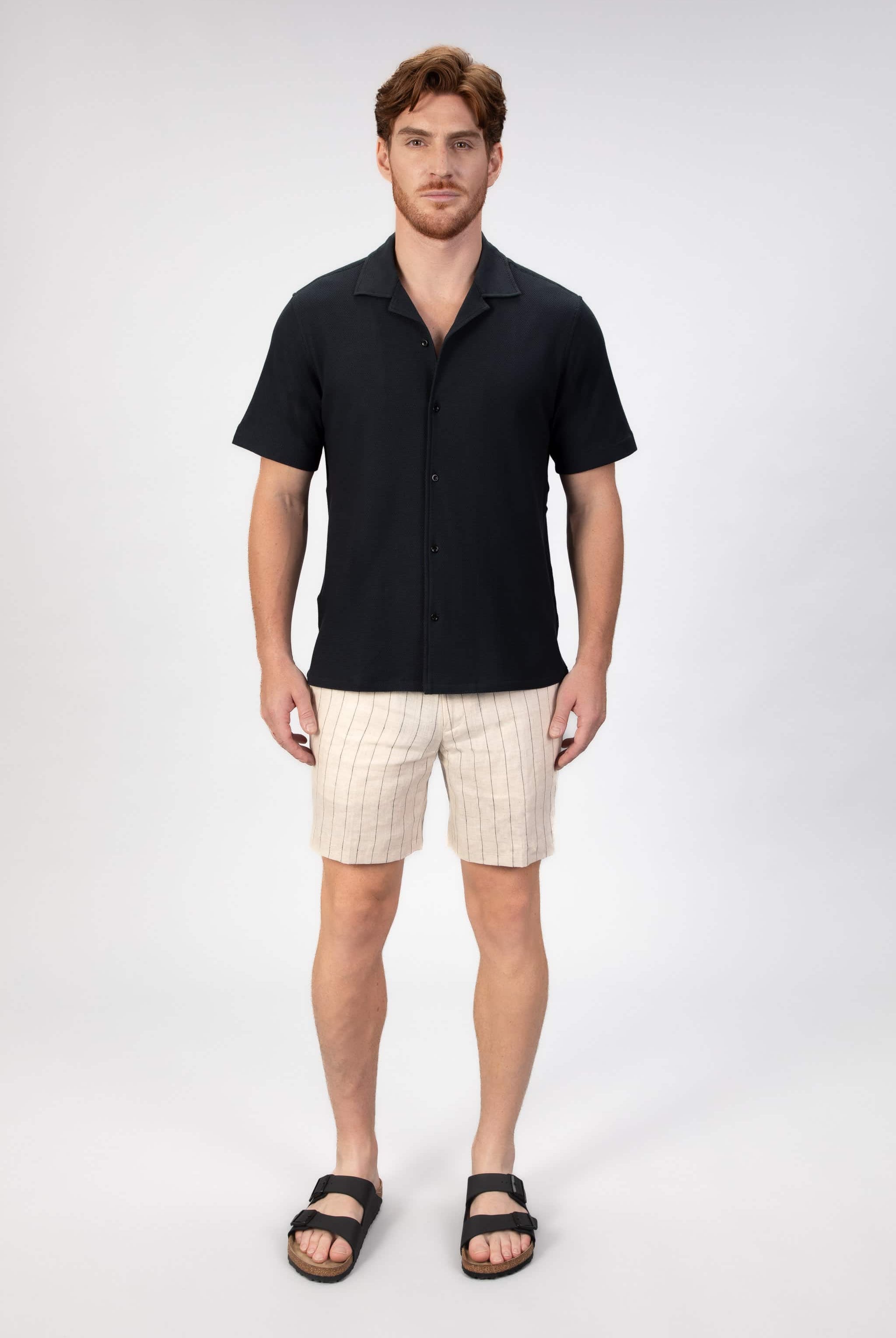 Man wearing a black short-sleeve camp collar shirt and beige stripe shorts on a plain background
