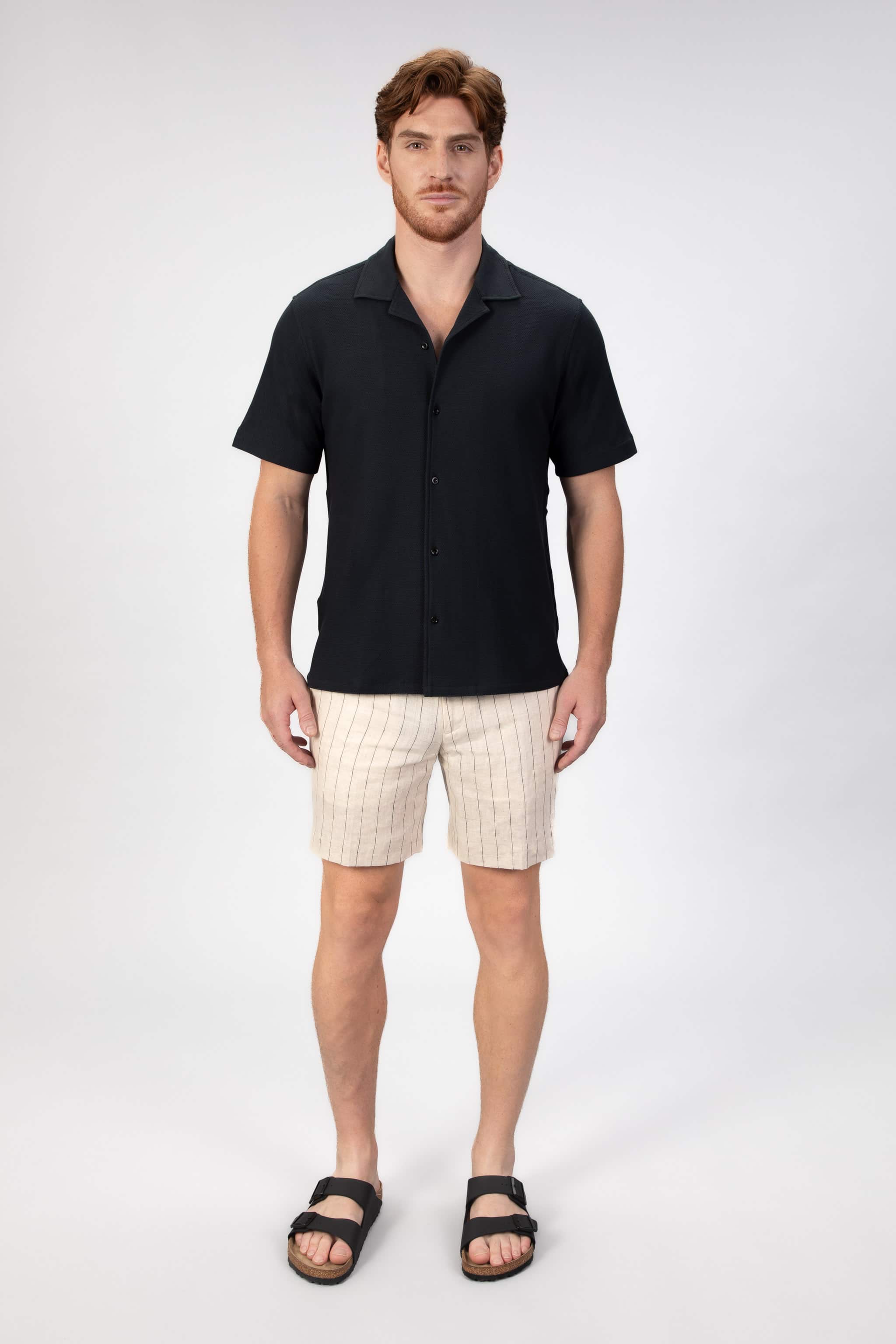 Man wearing a black short-sleeve camp collar shirt and beige stripe shorts on a plain background