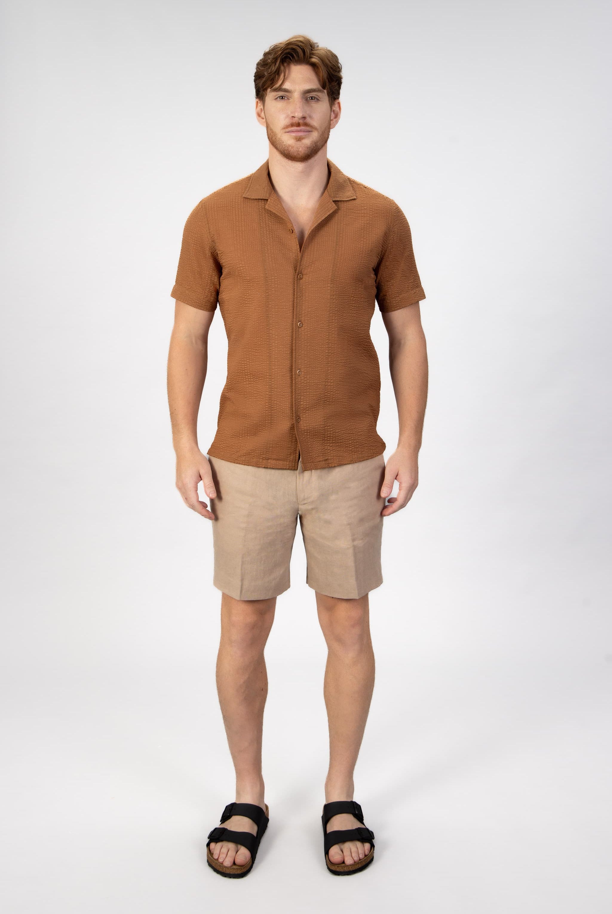 Man wearing a brown seersucker shirt and beige linen shorts on a light gray background