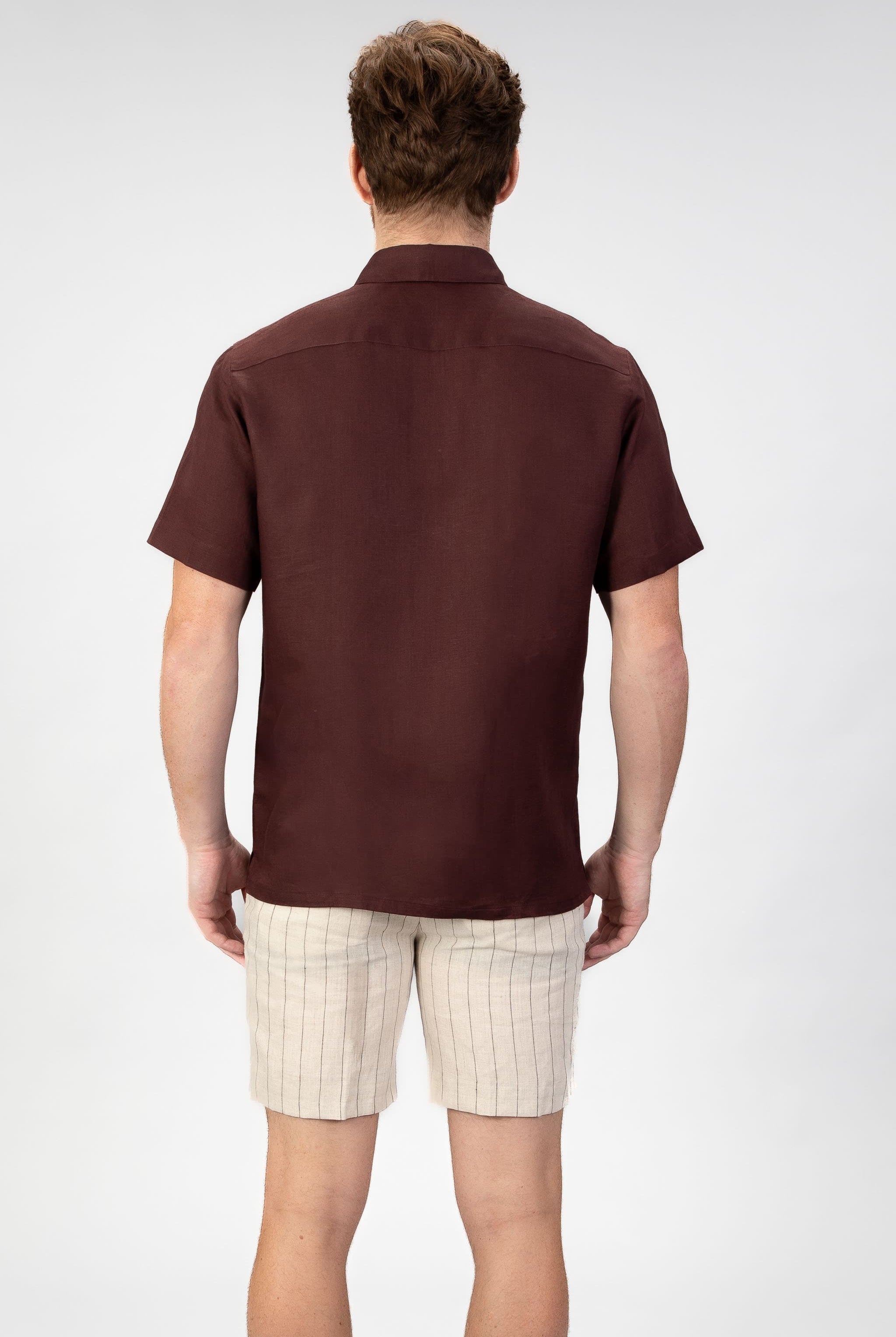 Man wearing a burgundy red short sleeve linen shirt with beige striped linen shorts on light gray background