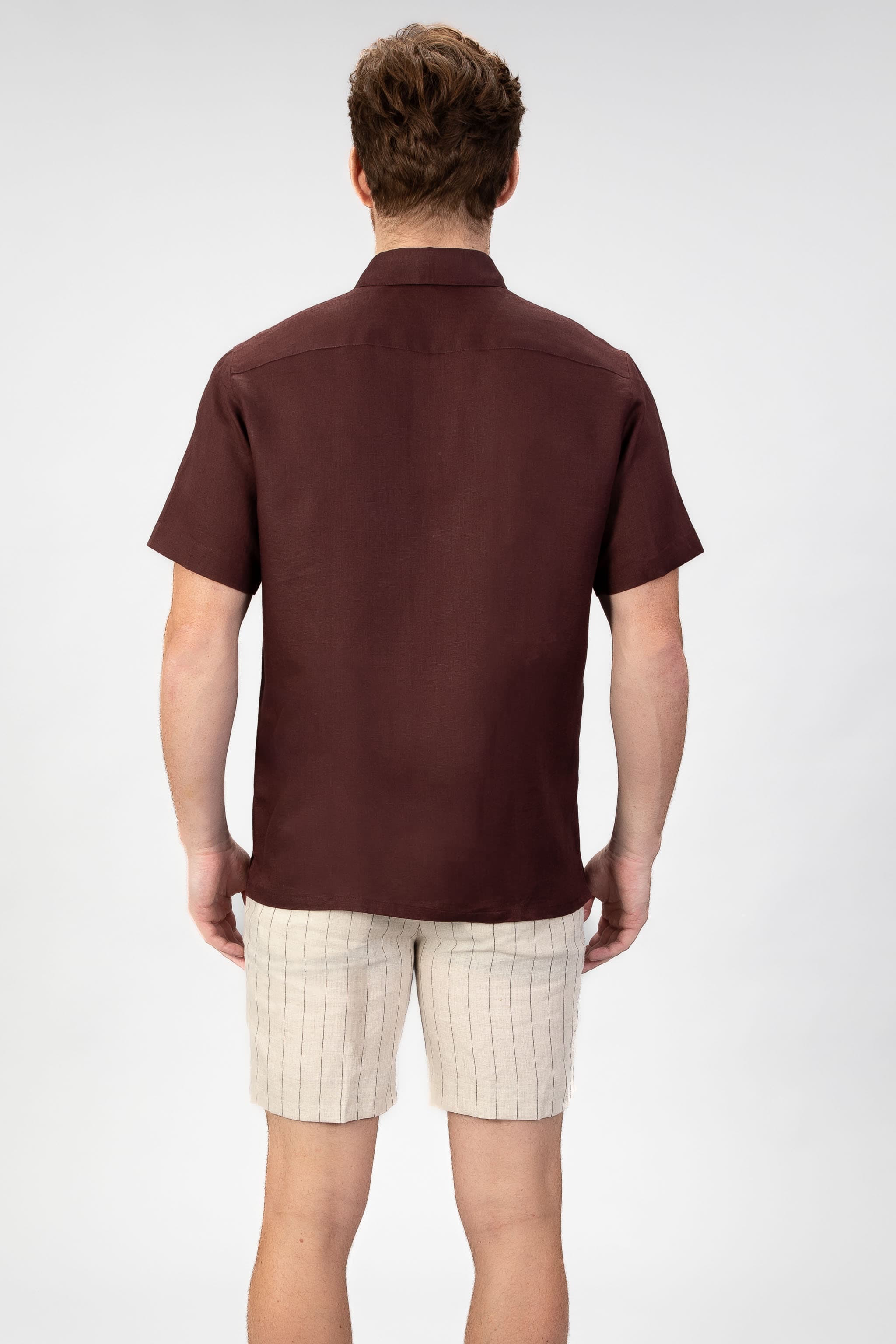 Man wearing a burgundy red short sleeve linen shirt with beige striped linen shorts on light gray background