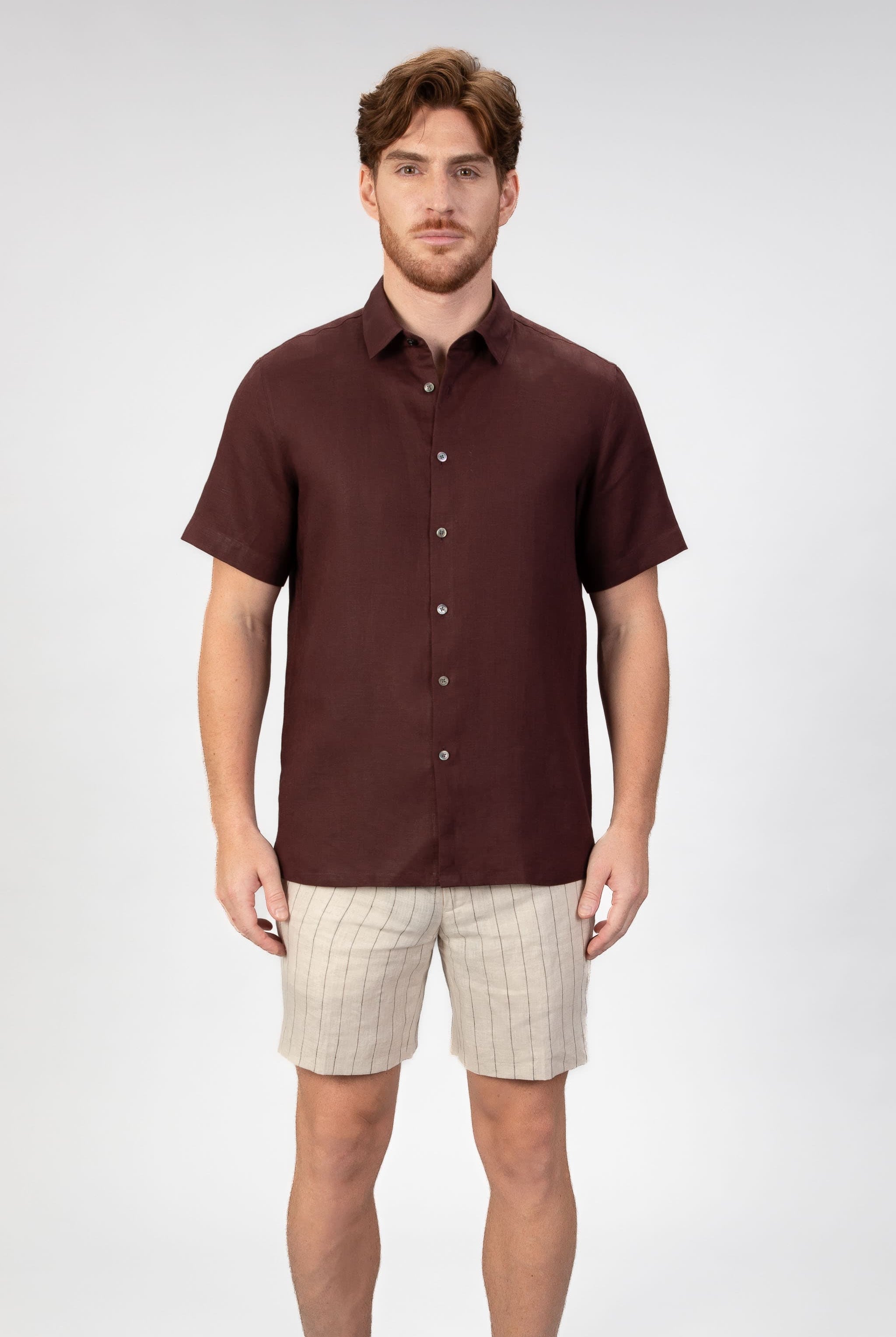 Man wearing a burgundy red short sleeve linen shirt with beige striped linen shorts on light gray background