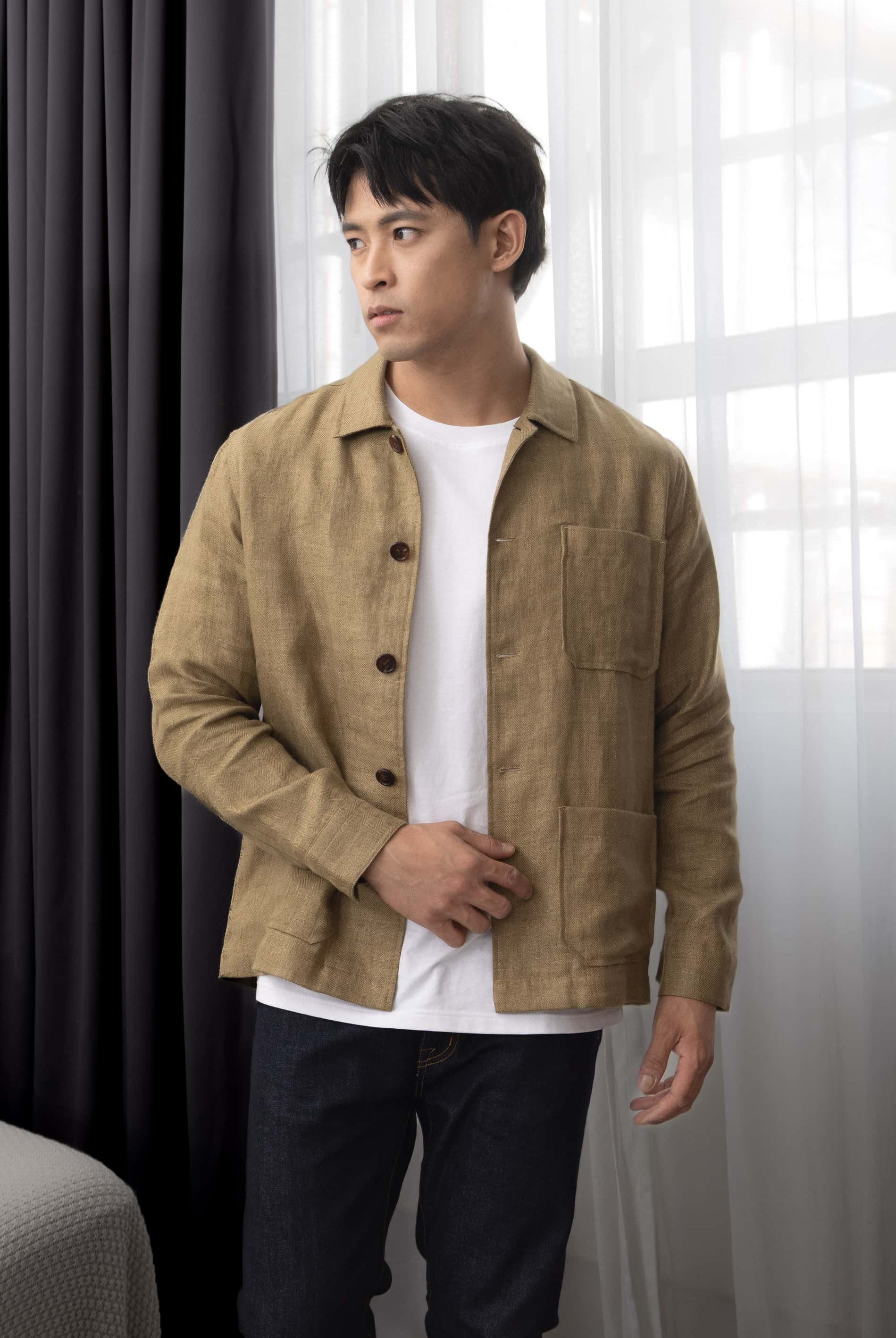 Man wearing a copper brown over-shirt over a white t-shirt, standing indoors with curtains in the background.