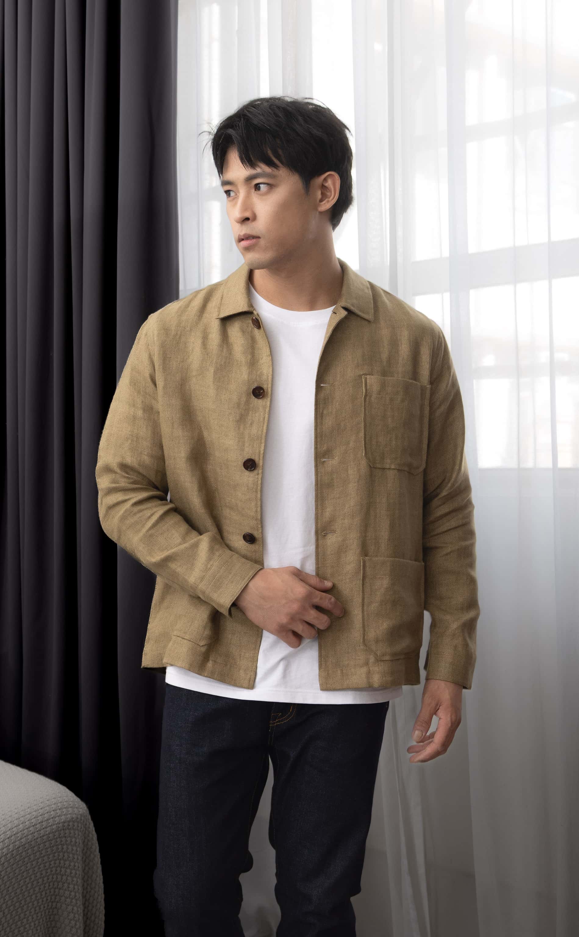 Man wearing a copper brown over-shirt over a white t-shirt, standing indoors with curtains in the background.