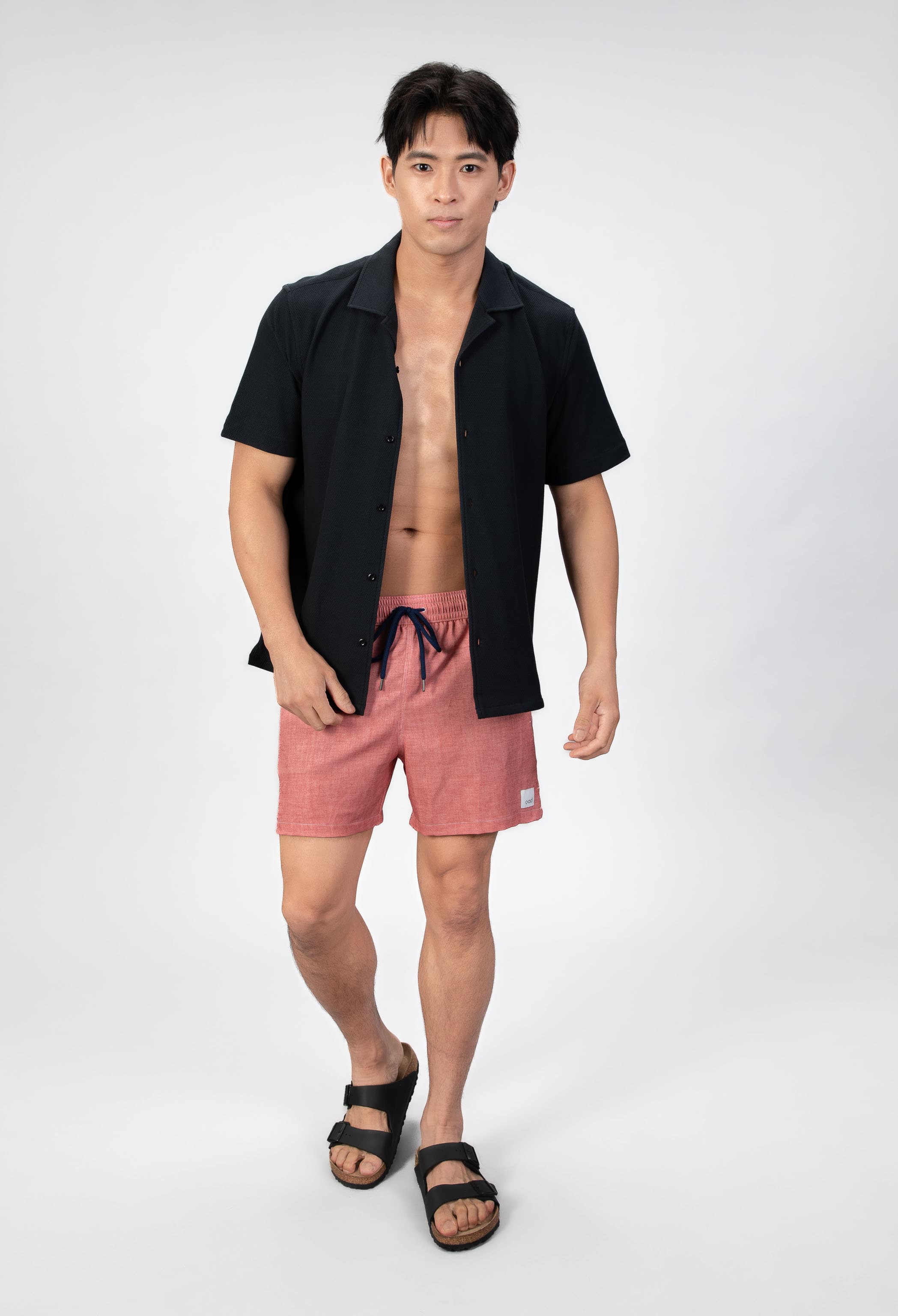 Person wearing pink-coral soft swim shorts and a black camp collar shirt on a light gray background