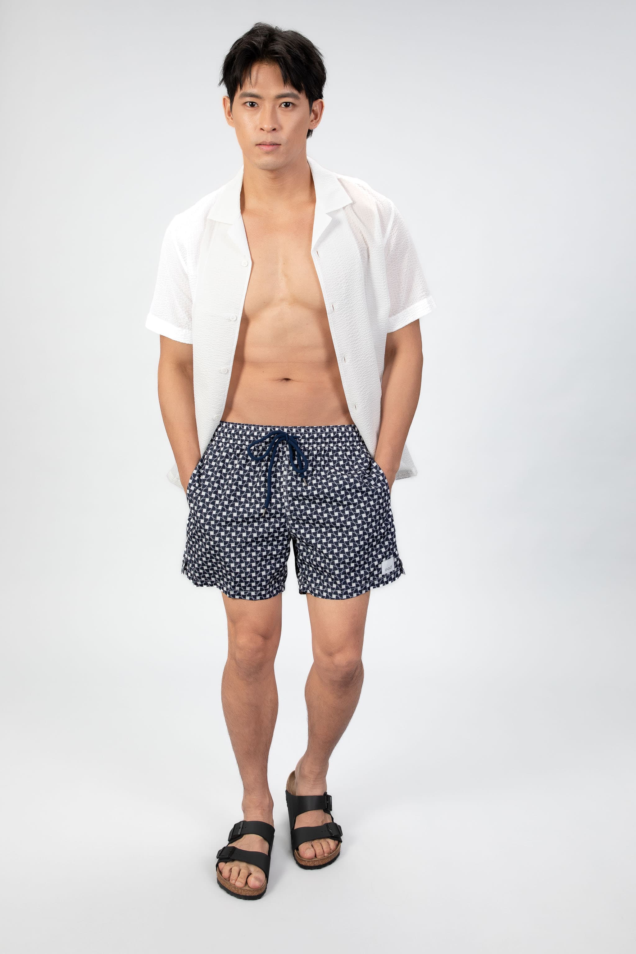 Front view of navy blue swim shorts with a subtle classic pattern and navy string waist closure. Paired with a thin white seersucker camp collar shirt.