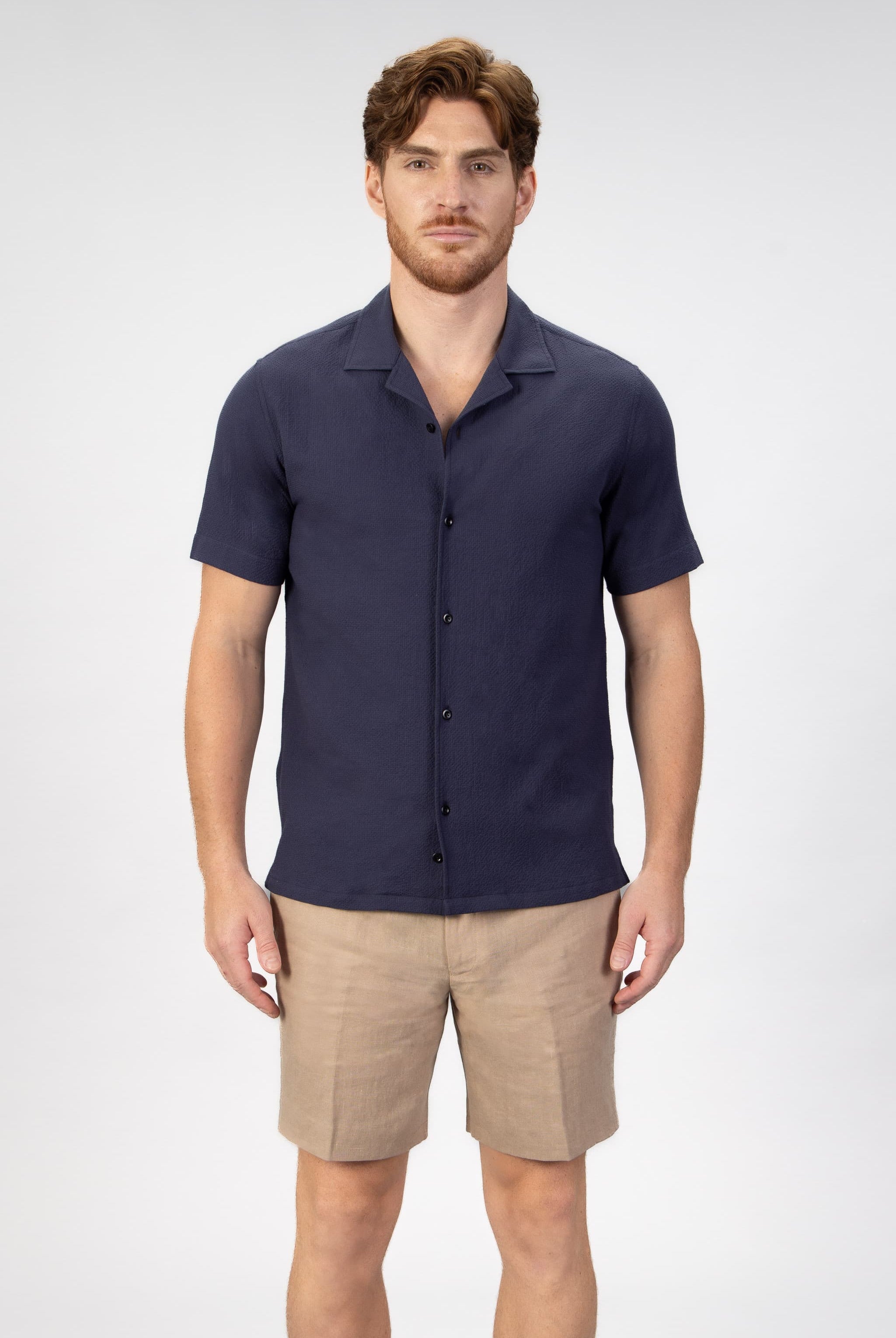 Man wearing a navy camp collar shirt sleeve shirt and beige shorts on a light gray background