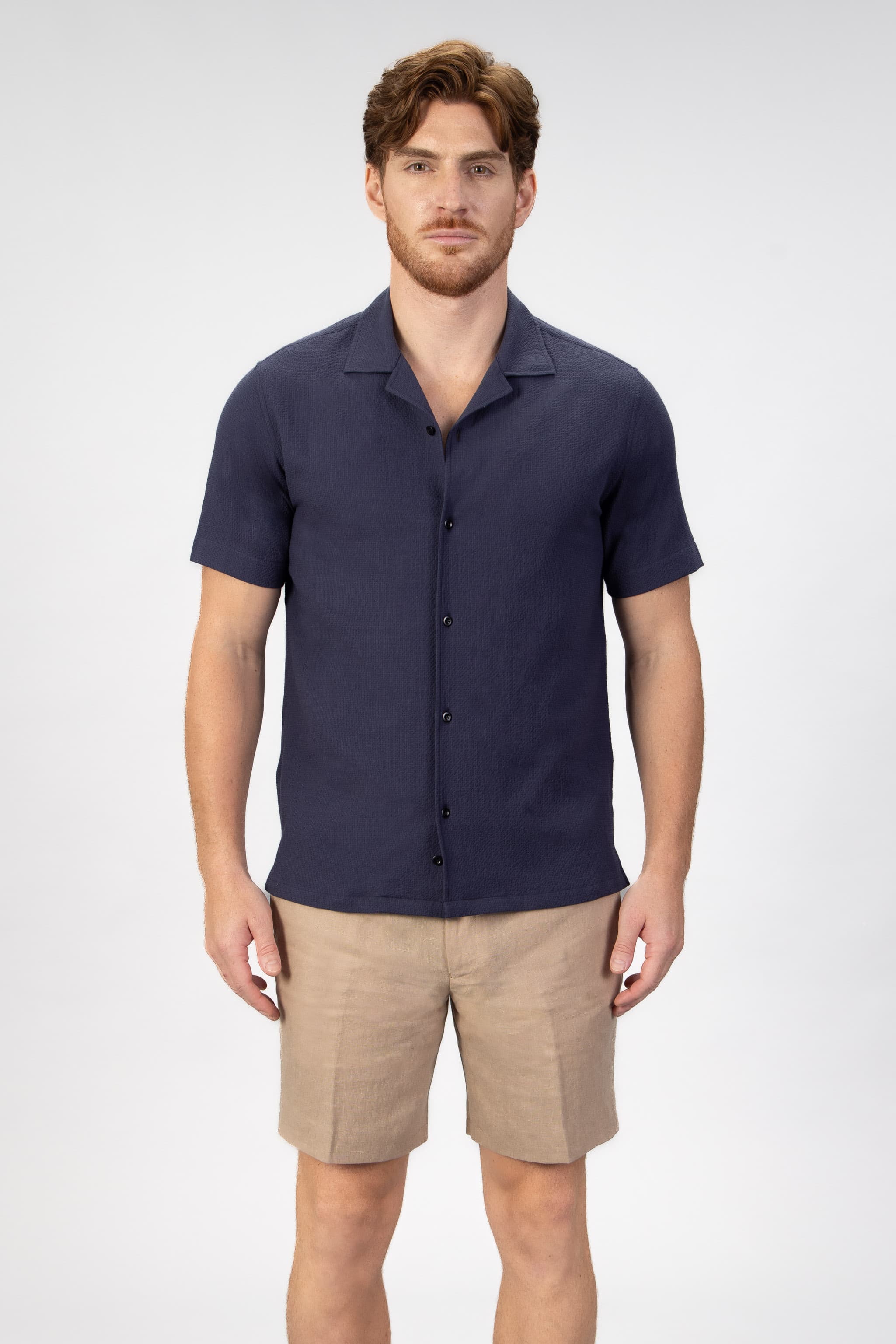 Man wearing a navy camp collar shirt sleeve shirt and beige shorts on a light gray background