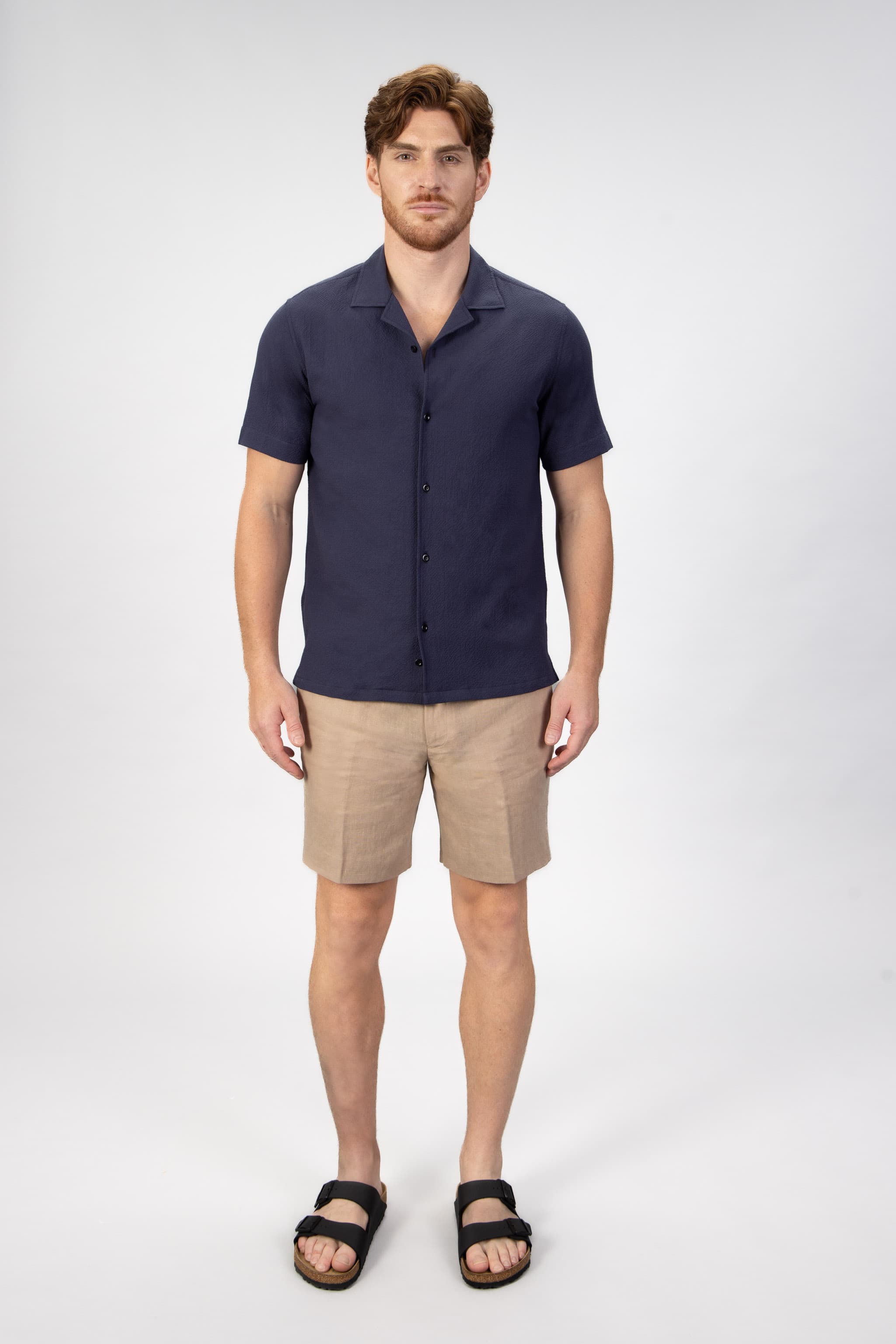 Man wearing a navy camp collar shirt sleeve shirt and beige shorts on a light gray background