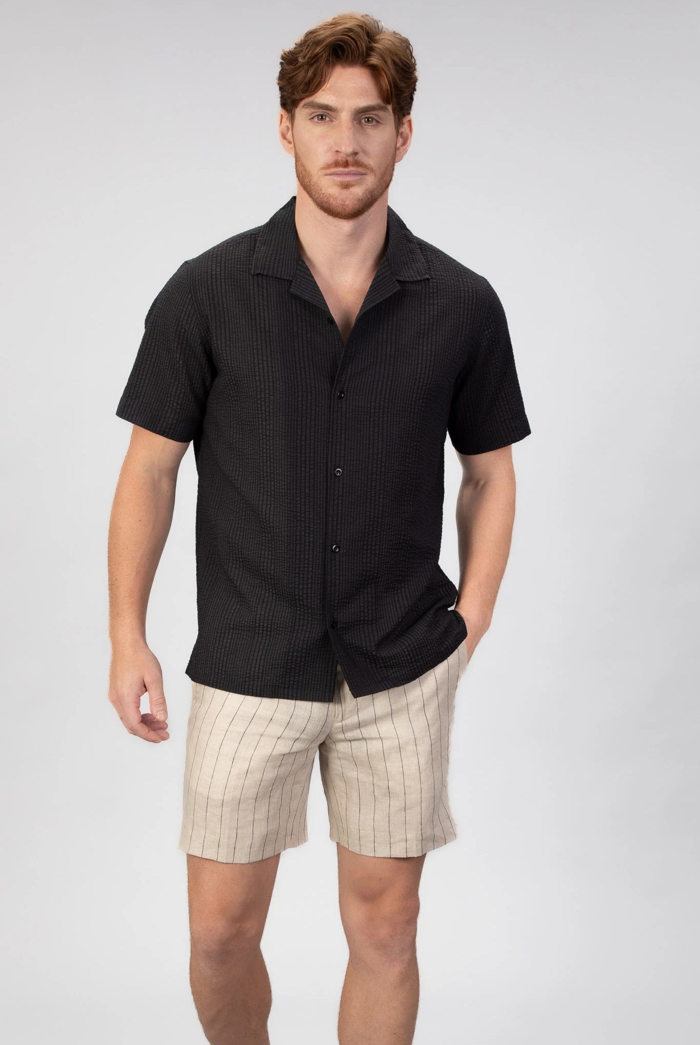 Man wearing a black short-sleeve camp collar shirt and beige shorts on a light gray background