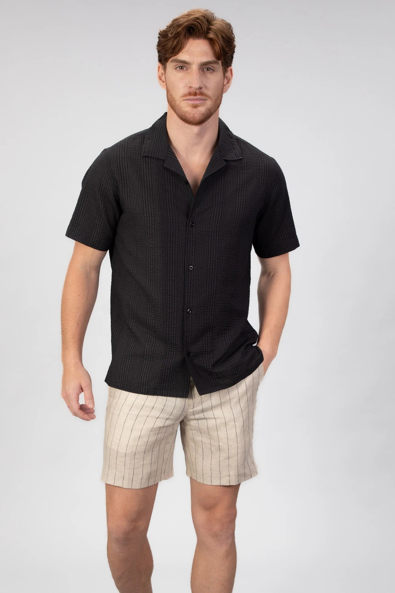 Man wearing a black short-sleeve camp collar shirt and beige shorts on a light gray background
