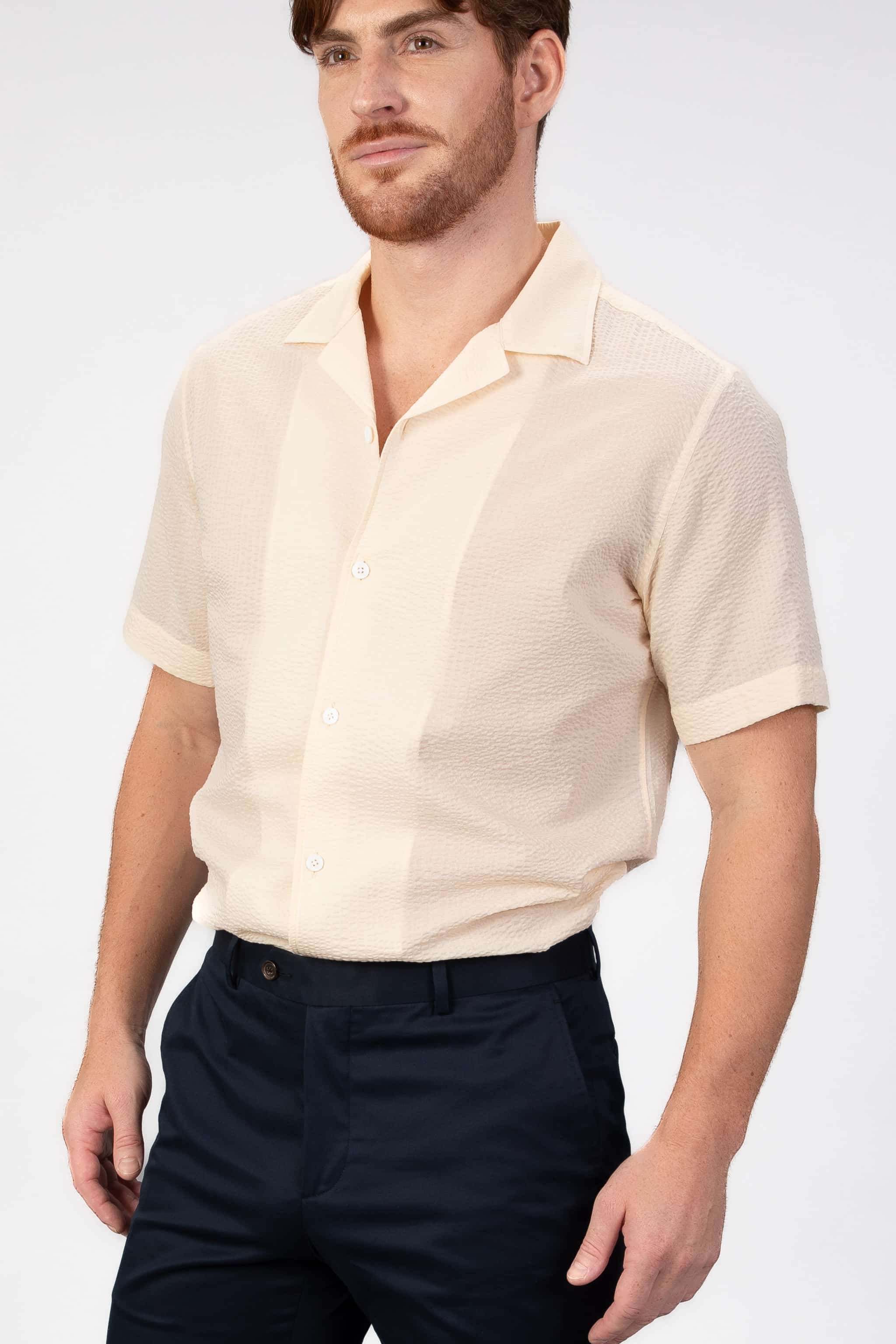 Man wearing a beige short-sleeve camp collar shirt and dark shorts on a white background