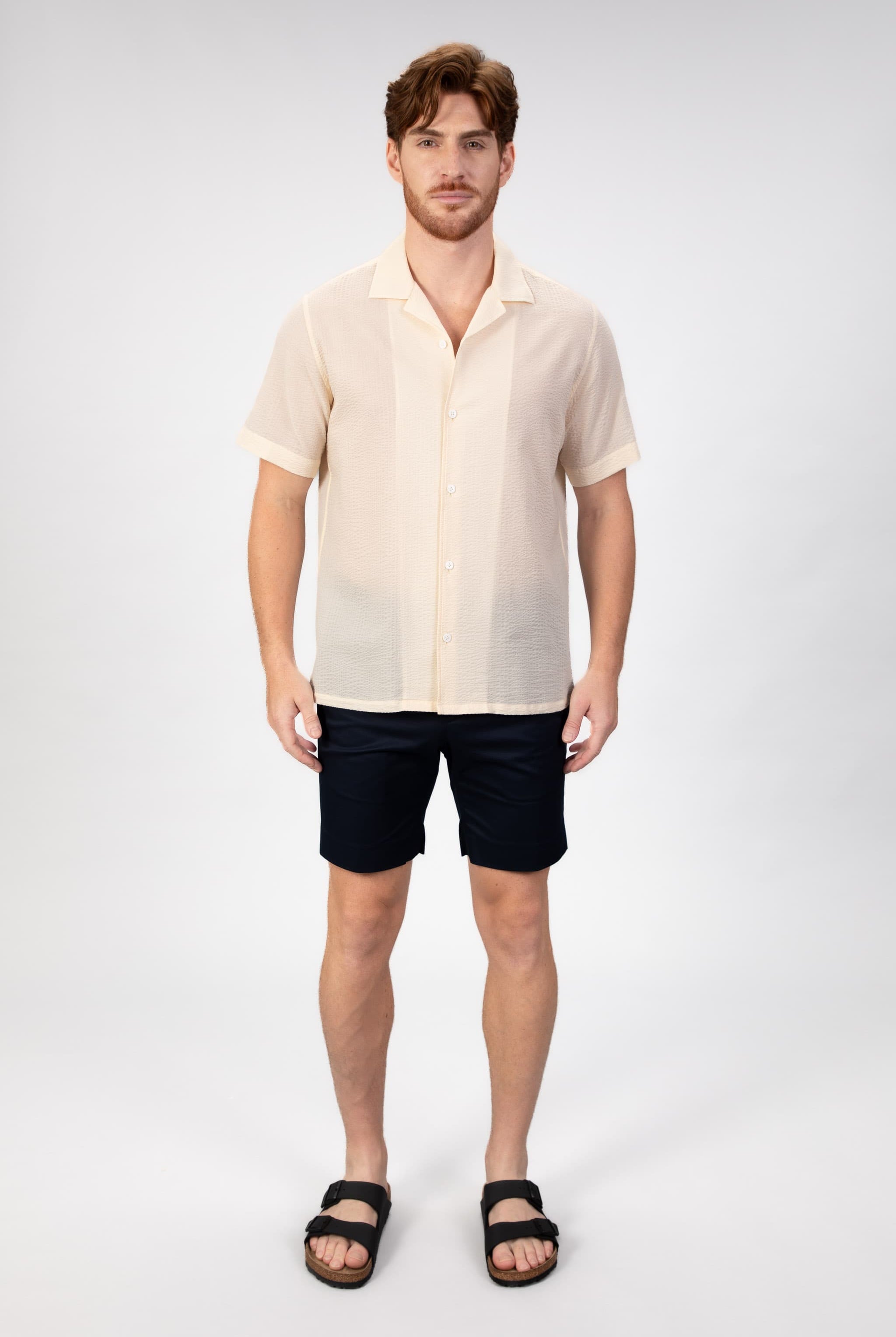 Man wearing a beige-sand seersucker camp collar shirt with navy blue cotton shorts on light gray background