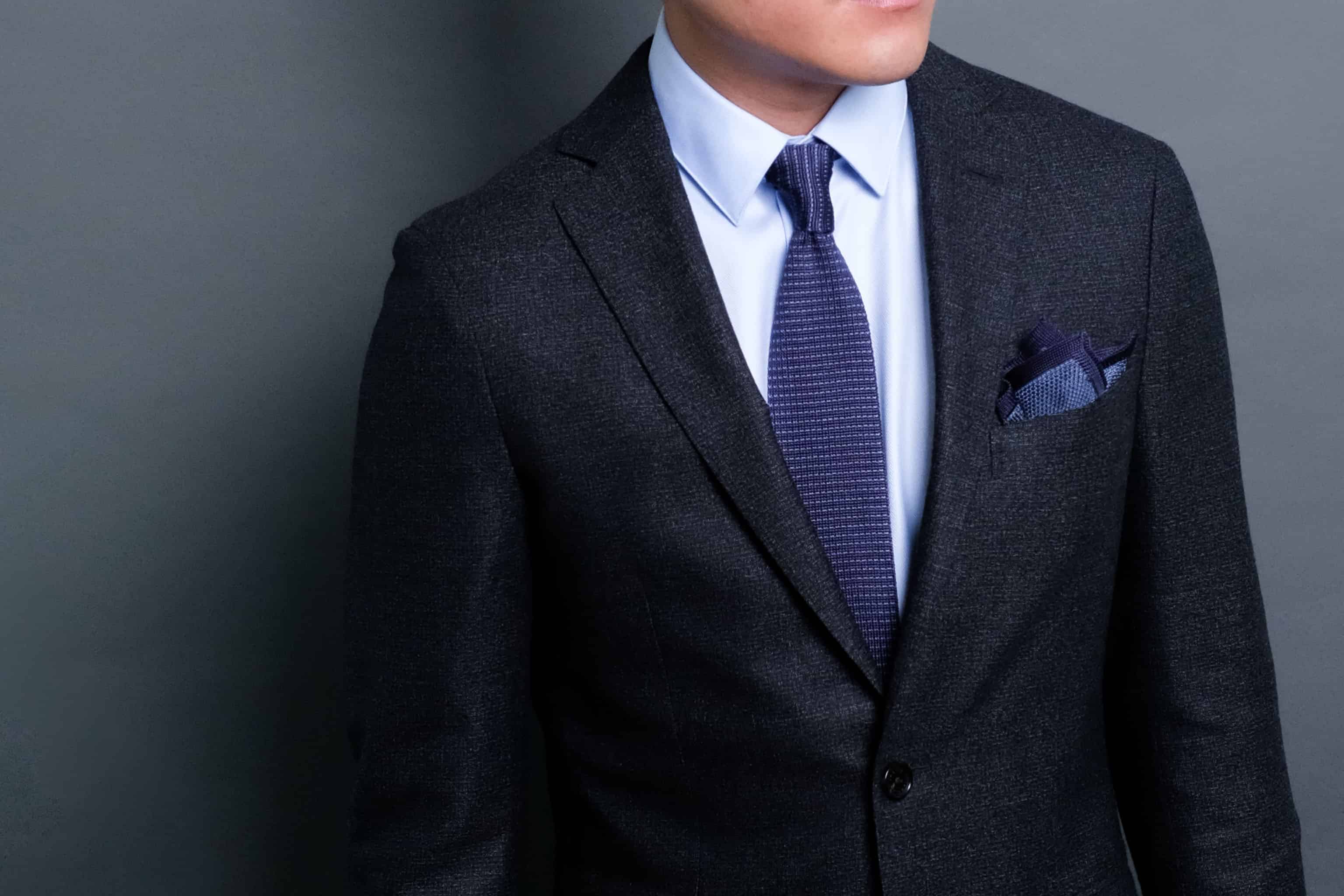 Man wearing a dark gray suit with a blue tie and pocket square against a gray background
