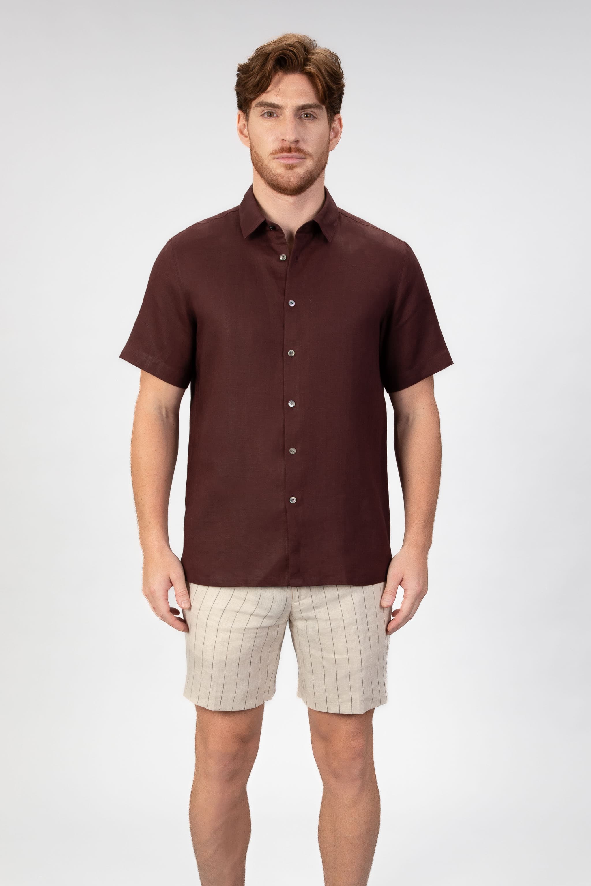Man wearing a burgundy red short sleeve linen shirt with beige striped linen shorts on light gray background