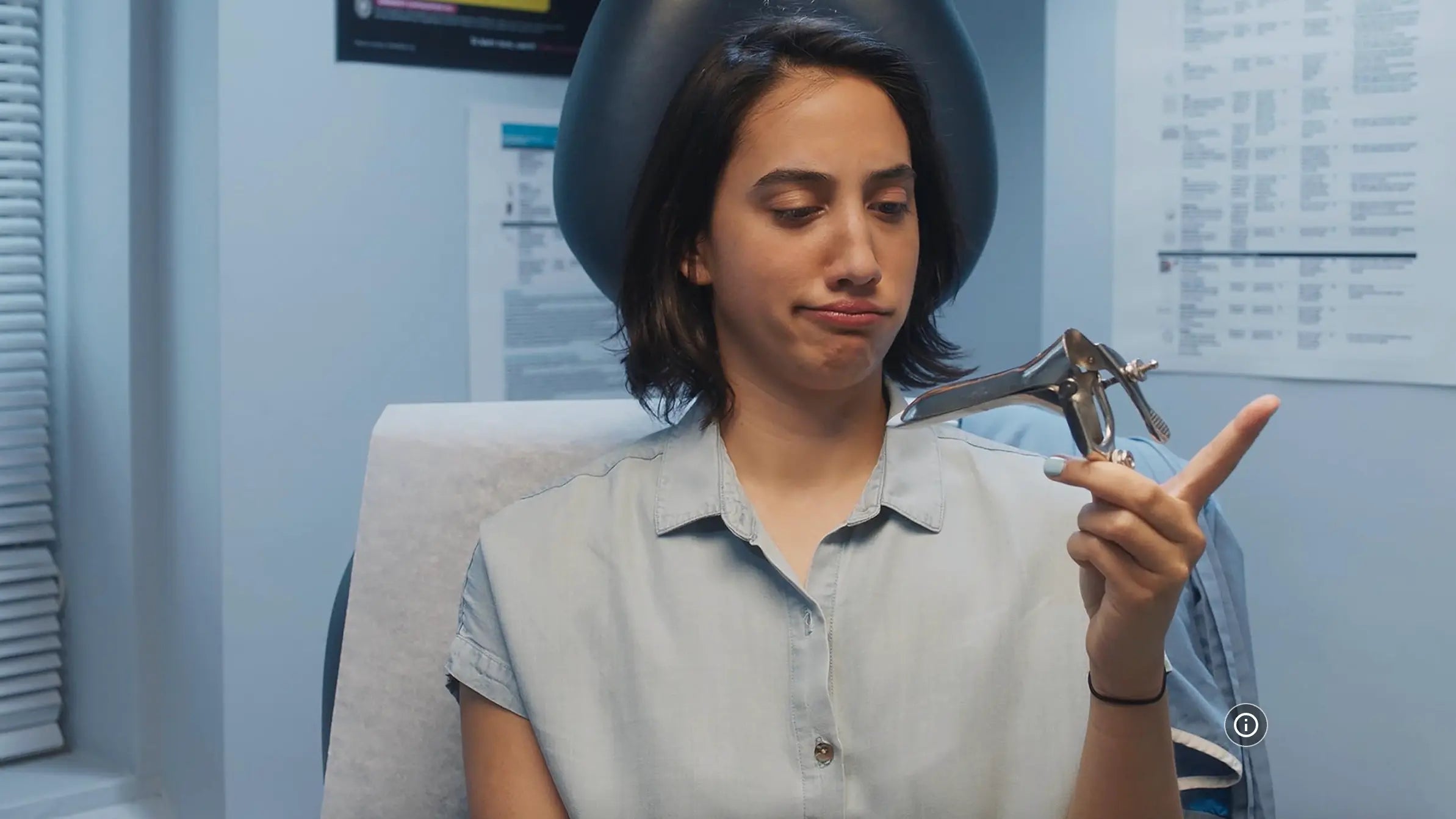 A woman in a light blue shirt sits in a dental chair holding a metal dental tool, with a dental office background visible.