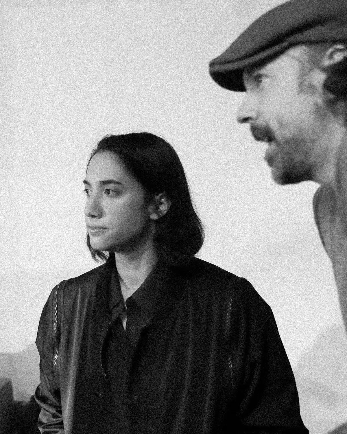 Black and white photograph featuring Julia Lindon in a dark jacket looking off-camera, with Jason Sudeikis wearing a flat cap partially visible on the right side.