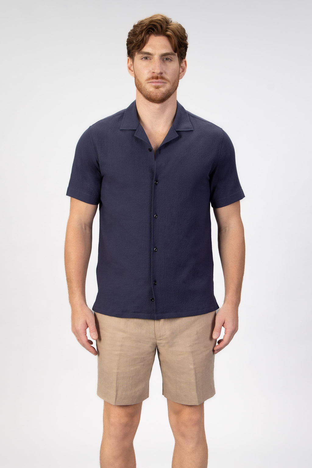 Man wearing a navy camp collar shirt sleeve shirt and beige shorts on a light gray background