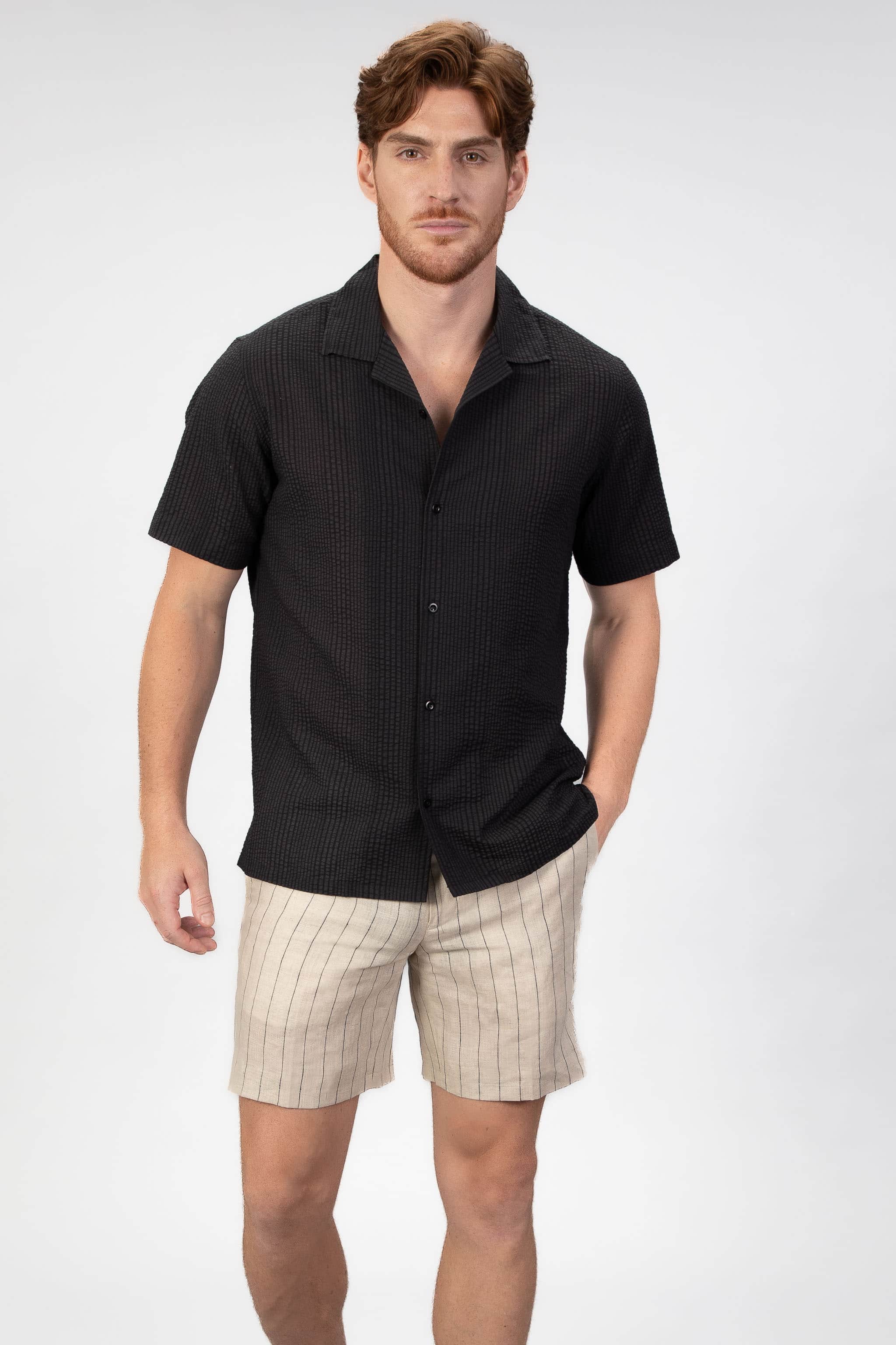 Man wearing a black short-sleeve shirt and beige shorts on a white background