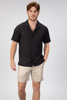 Man wearing a black short-sleeve camp collar shirt and beige shorts on a light gray background