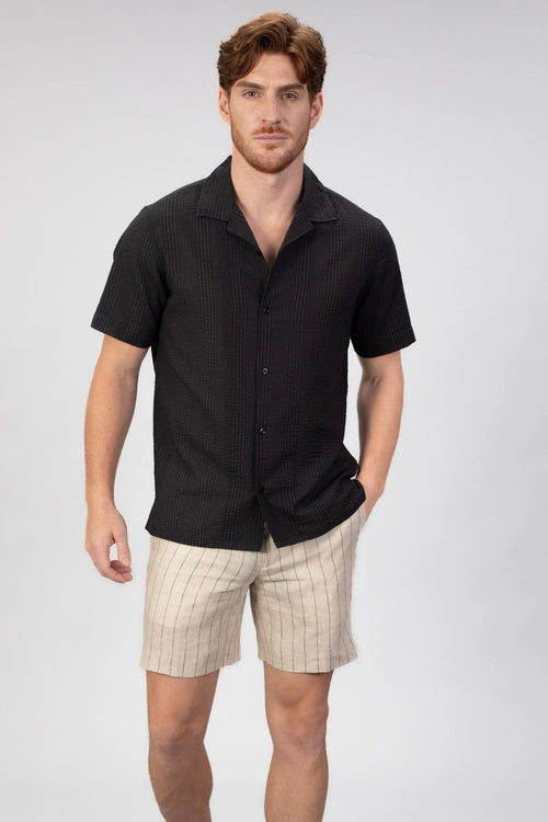 Man wearing a black short-sleeve camp collar shirt and beige shorts on a light gray background