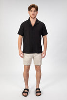 Man wearing a short sleeve camp collar shirt in black seersucker fabric with matching striped linen shorts in beige.