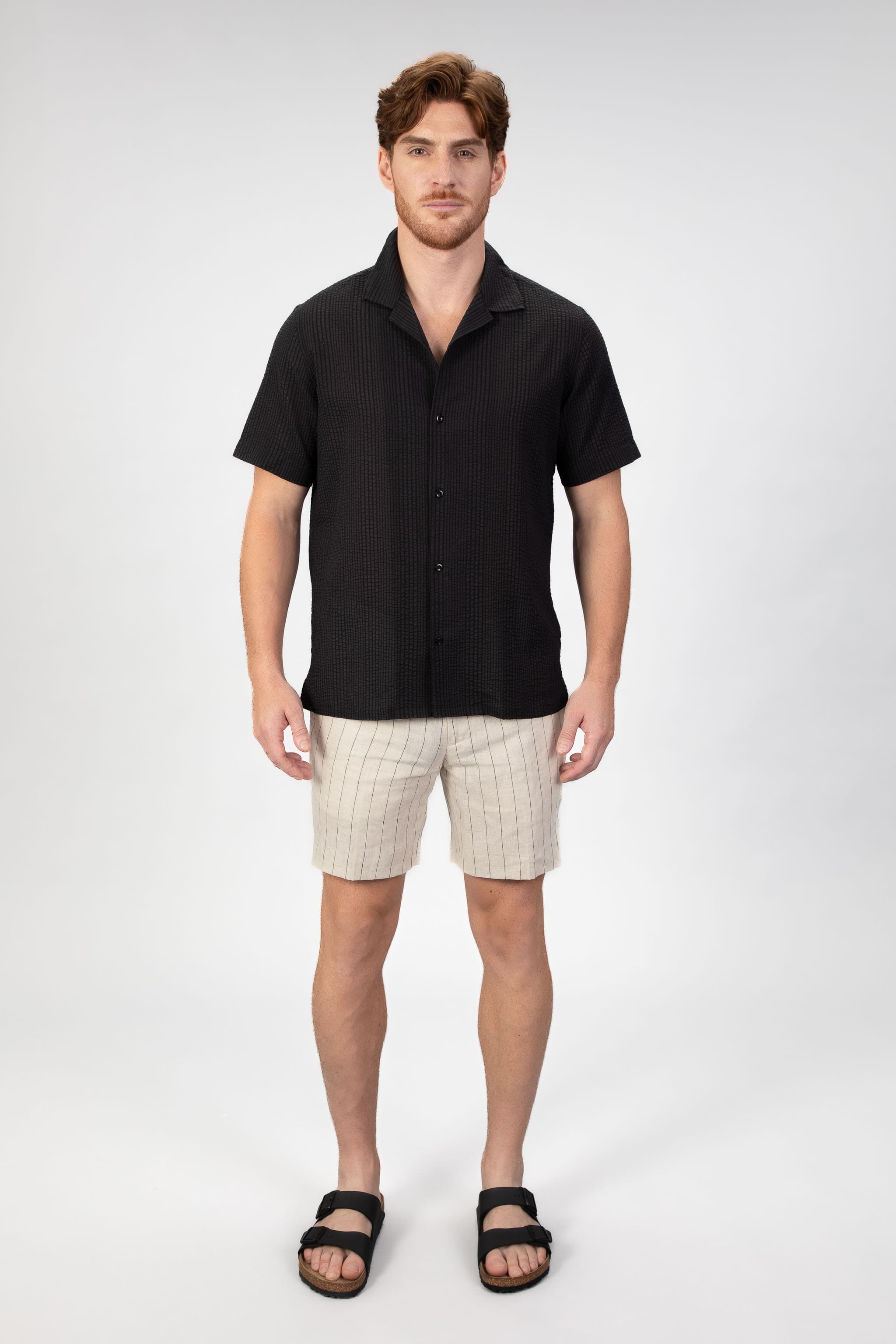 Man wearing a short sleeve camp collar shirt in black seersucker fabric with matching striped linen shorts in beige.