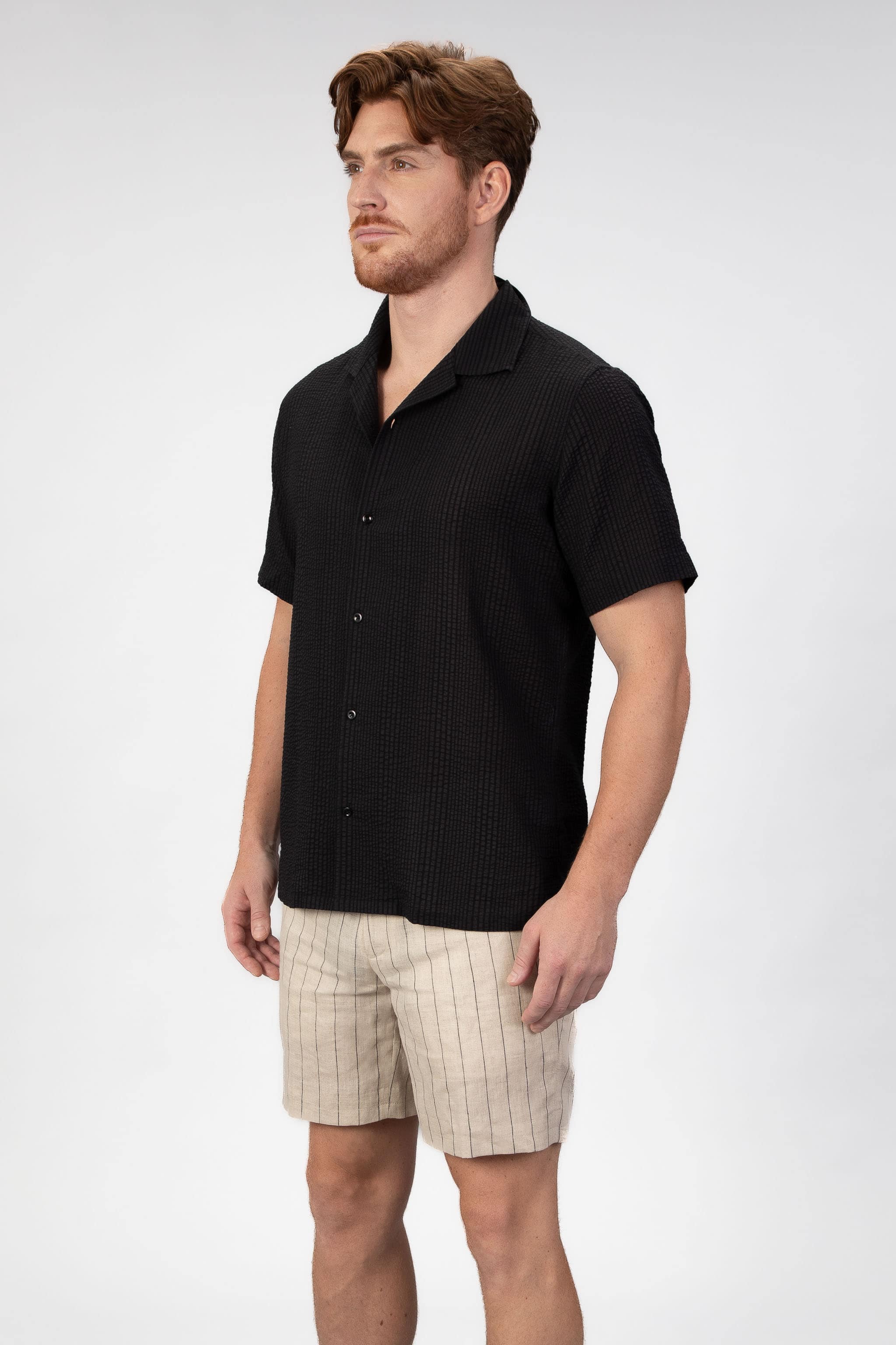 Man wearing a short sleeve camp collar shirt in black seersucker fabric with matching striped linen shorts in beige.
