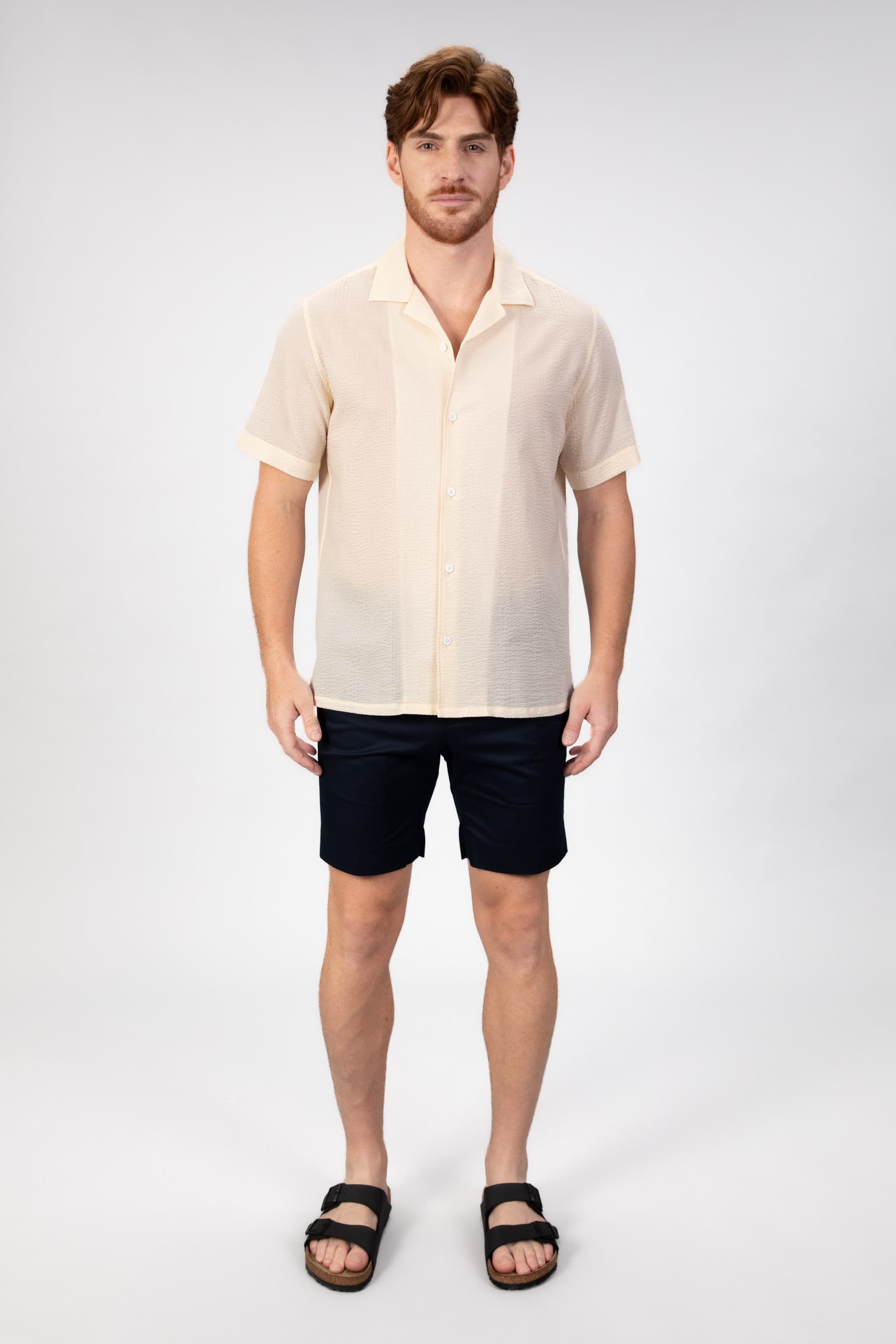 Man wearing a beige-sand seersucker camp collar shirt with navy blue cotton shorts on light gray background