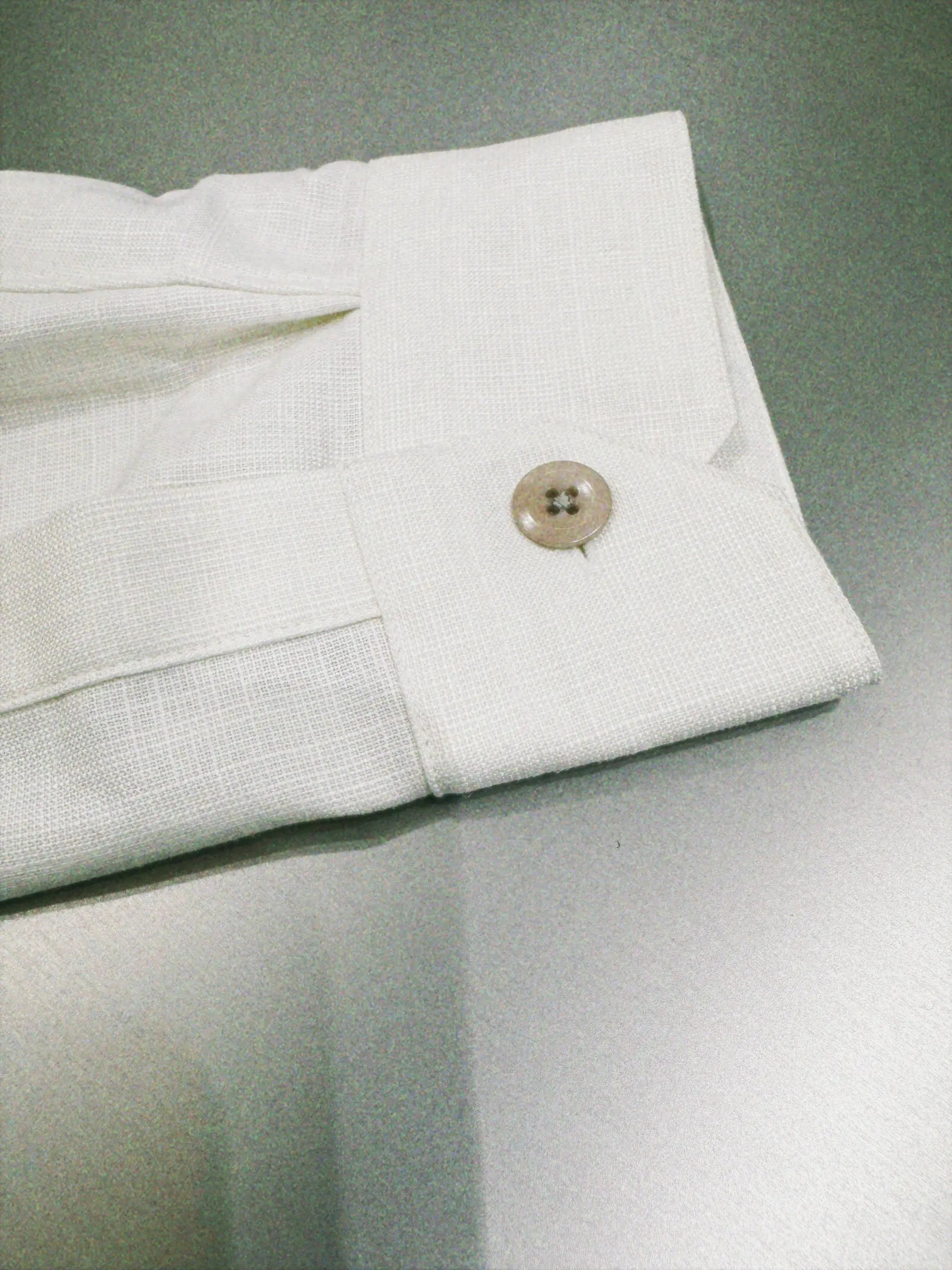 A close-up of a cream white cuff,silk, bamboo, wool linen shirt showing the fabric texture and a single light brown button on the cuff, set against a gray background.