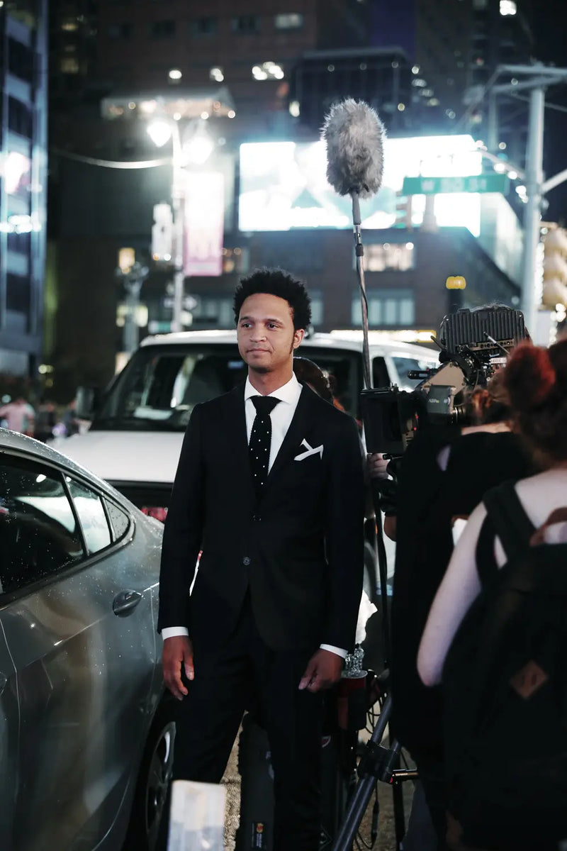 A man in a black suit, white shirt, and black tie stands on a city street at night, surrounded by cameras, a microphone, and other people in a paparazzi-style scene.