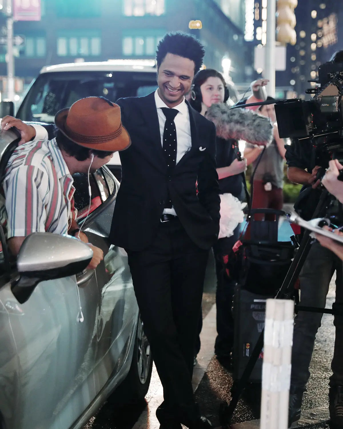 Chip Hamilton in a navy once a day suit and tie walking outdoors at night near a car, surrounded by photographers and people in a city setting.