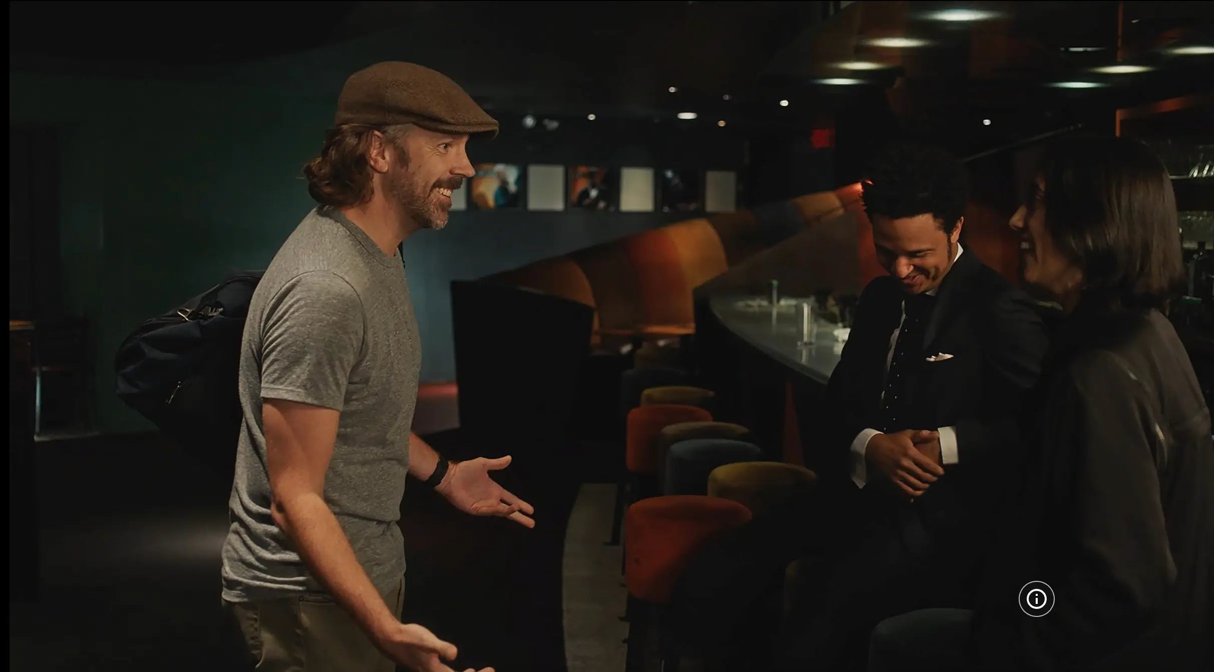 Jason Sudeikis in gray t-shirt and brown cap gestures while talking to two people seated at a bar in a dimly lit restaurant or lounge. from lady liberty film 2019