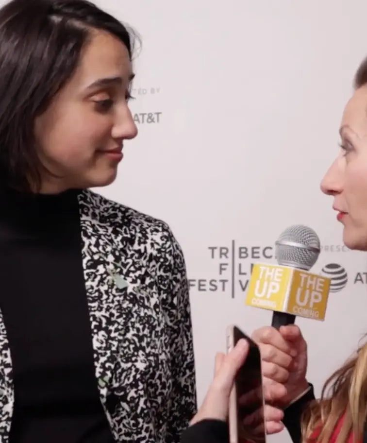 A woman in a black and white patterned blazer over a black turtleneck is being interviewed by a reporter holding a microphone with a yellow 'The Up' logo, set against a white Tribeca Film Festival backdrop.