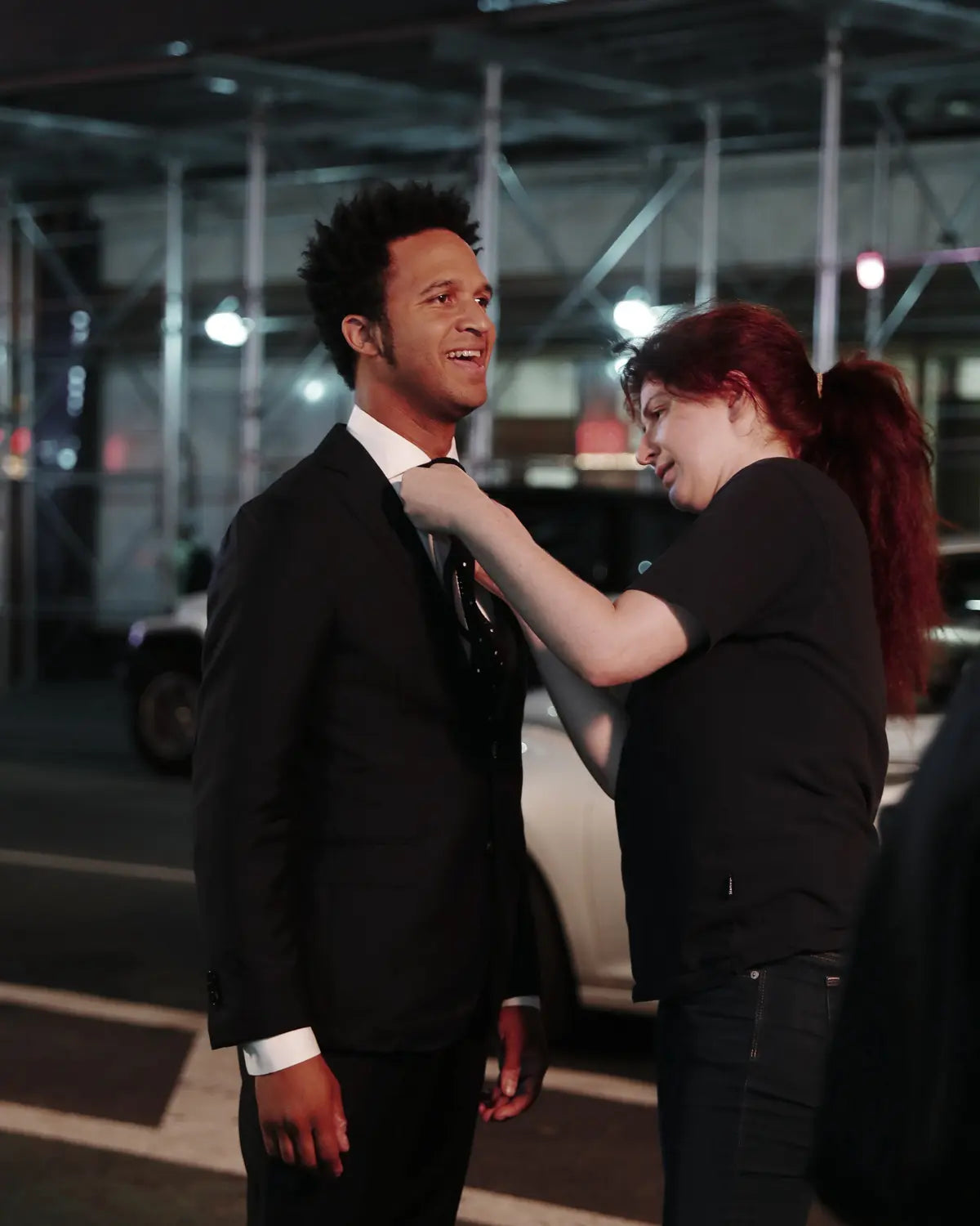 A man in a dark navy suit and white shirt stands while a woman in a black T-shirt adjusts his tie, photographed at night in an urban setting with scaffolding and street elements in the background.