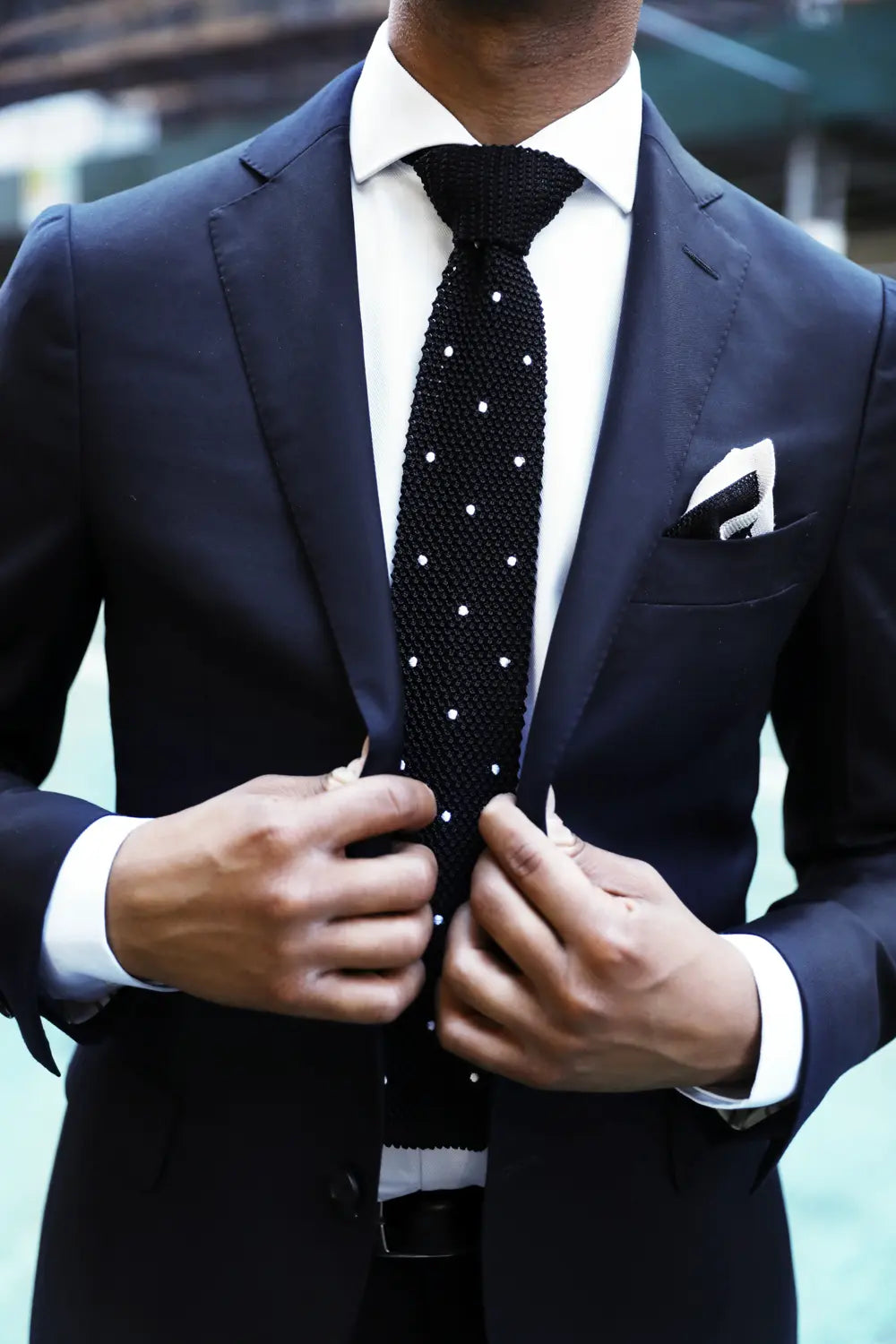 A close-up of a man wearing a navy blue suit jacket, white dress shirt, and a black textured tie with white polka dots. He is adjusting the front of the jacket, with a white and black knitted pocket square in his breast pocket.