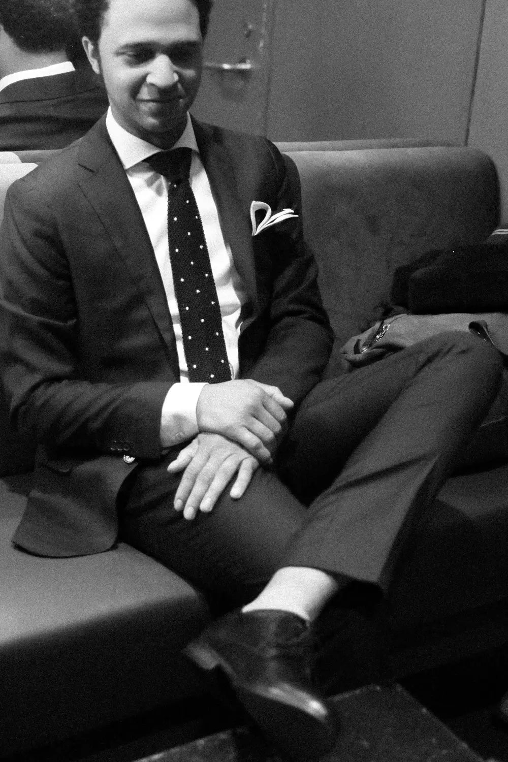 A man wearing a navy suit, white dress shirt, and a polka-dot tie, sitting on a dark leather couch in a dimly lit room. Black and white image