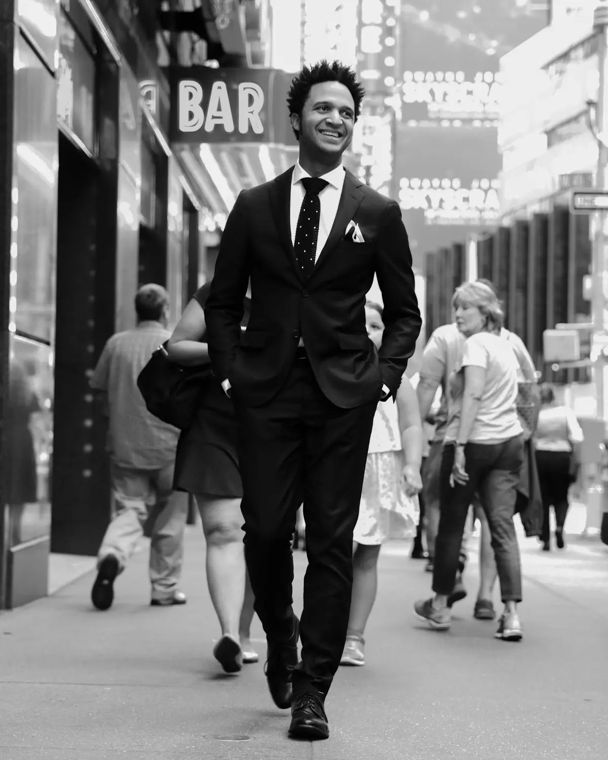 black and white image, A man in a dark suit, white shirt, and patterned tie walks down a city street in black and white, with blurred pedestrians and storefront signs in the background.
