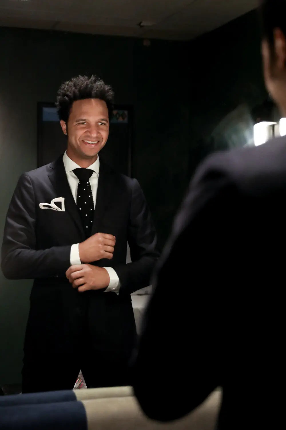 A man in a black suit, white dress shirt, and black polka-dot tie stands in front of a mirror, smiling. The foreground shows the back of another person's head and shoulder, and the background is a dark indoor room.